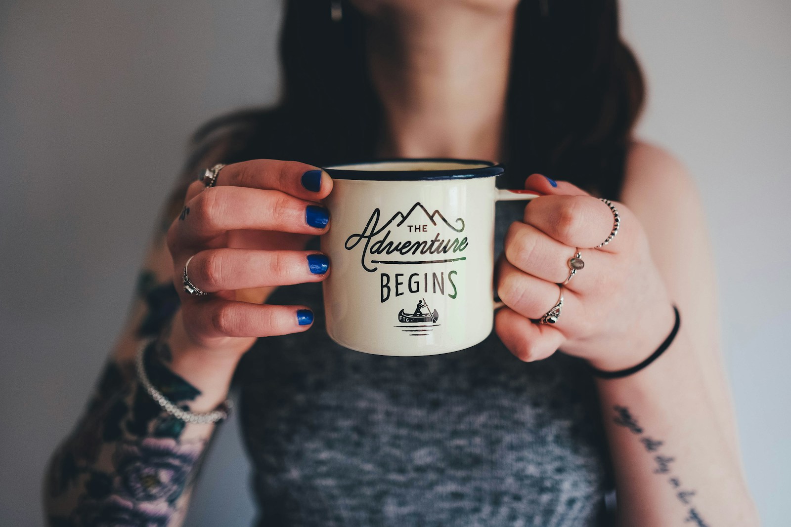 woman with tattoo on arms holding a coffee mug that says the adventure begins