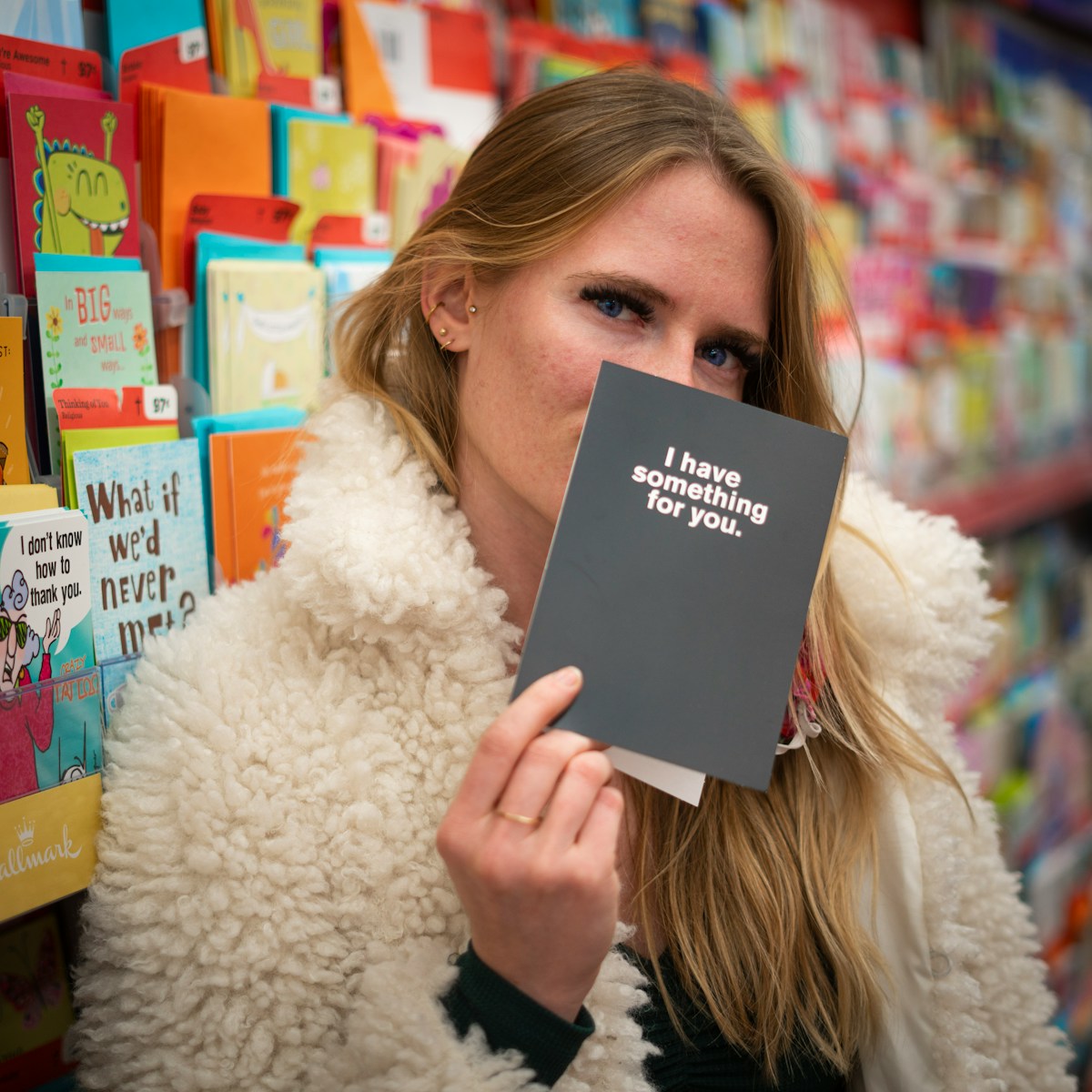 woman in a white fur coat holding a card with other greeting cards in the background. the card that she is holding says i have something for you